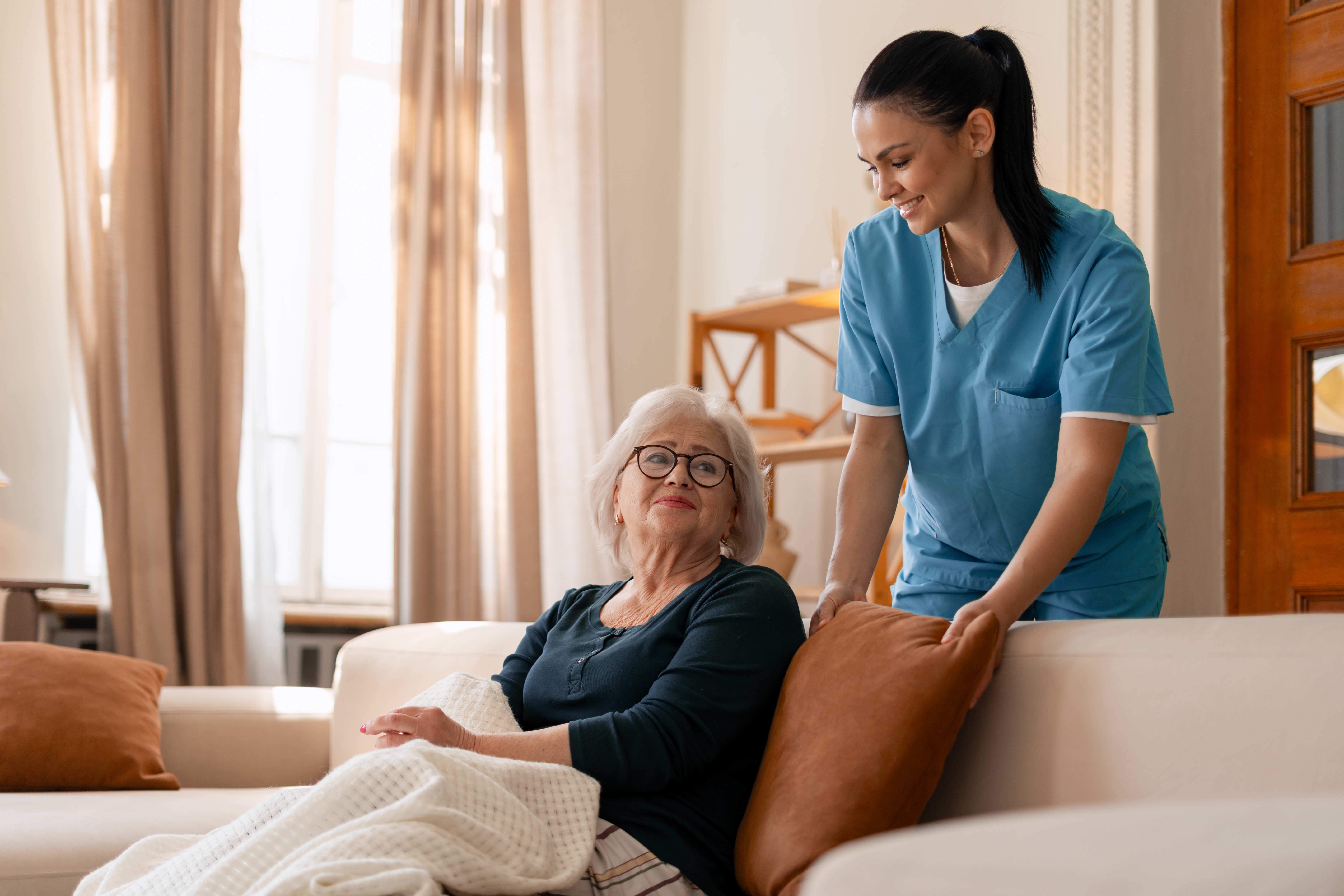 Professional caregiver helping a senior woman sitting on a sofa at home, providing comfortable cushions and friendly service